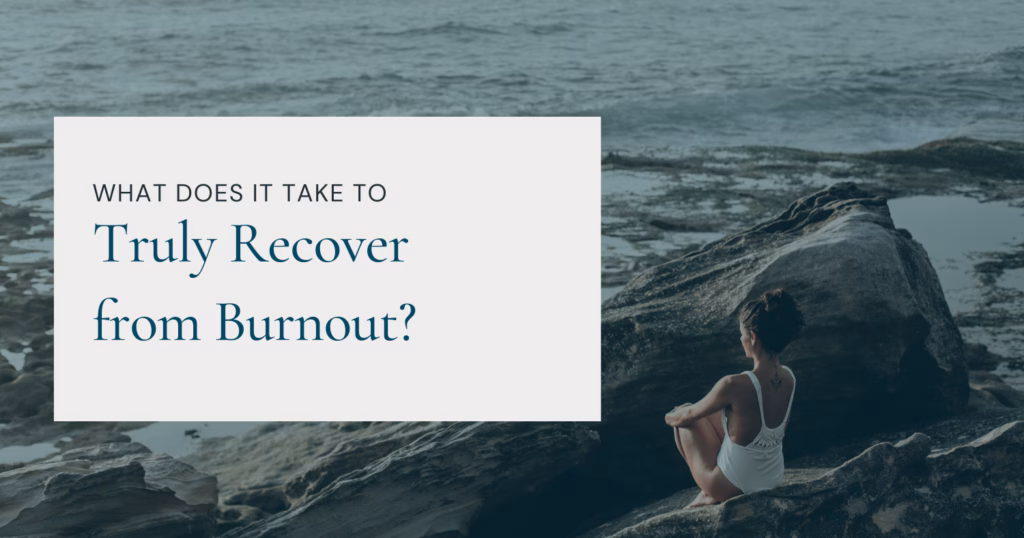 A woman in a white swimsuit sits on a rocky shoreline, gazing out at the ocean in a moment of solitude and reflection. The overlaid text reads, 'What does it take to truly recover from burnout?' The serene, natural setting emphasizes relaxation and self-care, reinforcing the theme of burnout recovery.